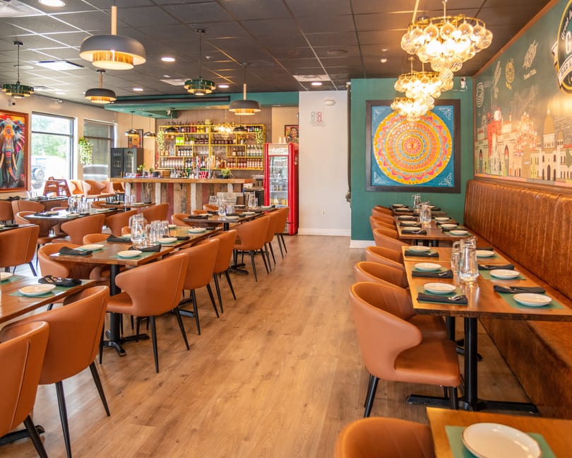 Spacious restaurant interior with set tables, a bar, and colorful Indian-inspired artwork on the walls.