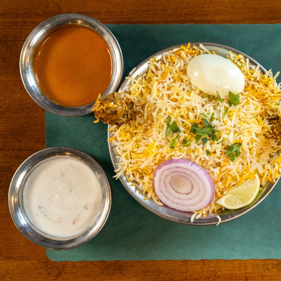 Chicken biryani with a boiled egg, onion, lime, and two side sauces on a green mat.