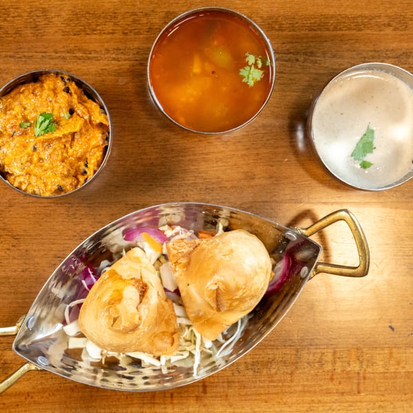 Two golden-brown samosas in a silver dish with shredded cabbage and red onions, alongside three bowls of sauces.