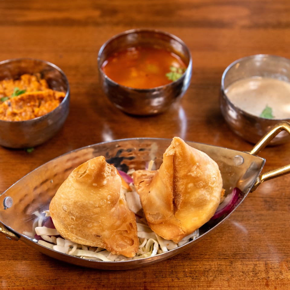 Two golden samosas on shredded cabbage in a metal dish, with three small bowls of sauces behind them.