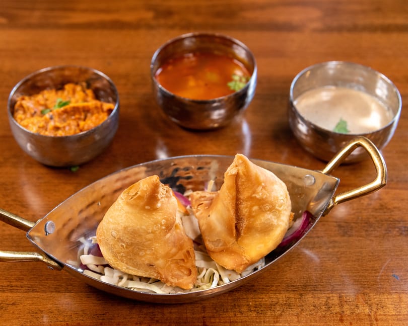 Two golden samosas on shredded cabbage in a metal dish, with three small bowls of sauces behind them.