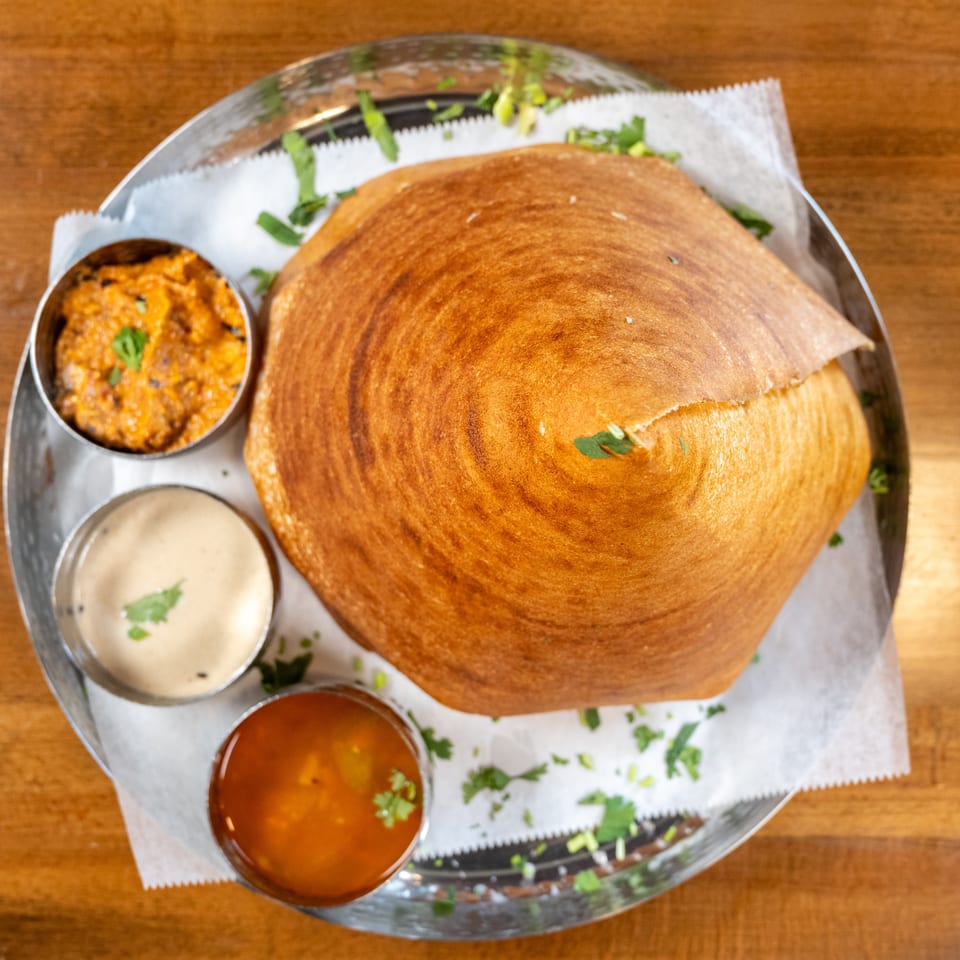 Crispy golden dosa served with three small bowls of colorful chutneys and sambar on a metal plate.