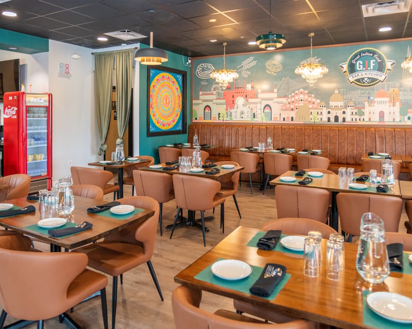 Interior of GIF Bar & Kitchen, an Indian restaurant, with empty tables, brown chairs, and a colorful mural.