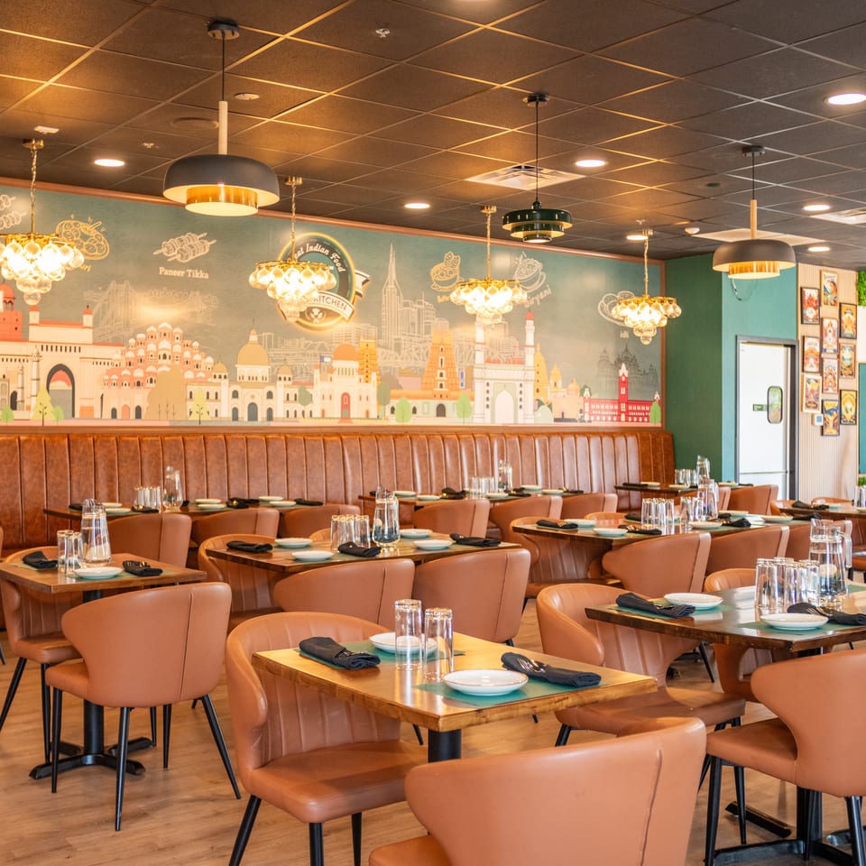 A modern Indian restaurant interior with brown leather chairs, wooden tables, and a mural of Indian landmarks.