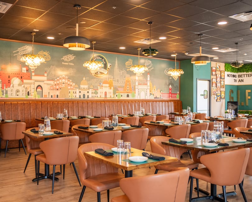 A modern Indian restaurant interior with brown leather chairs, wooden tables, and a mural of Indian landmarks.