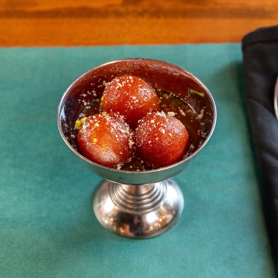 Three gulab jamun in a silver cup, garnished with powdered sugar and pistachios, on a green placemat.