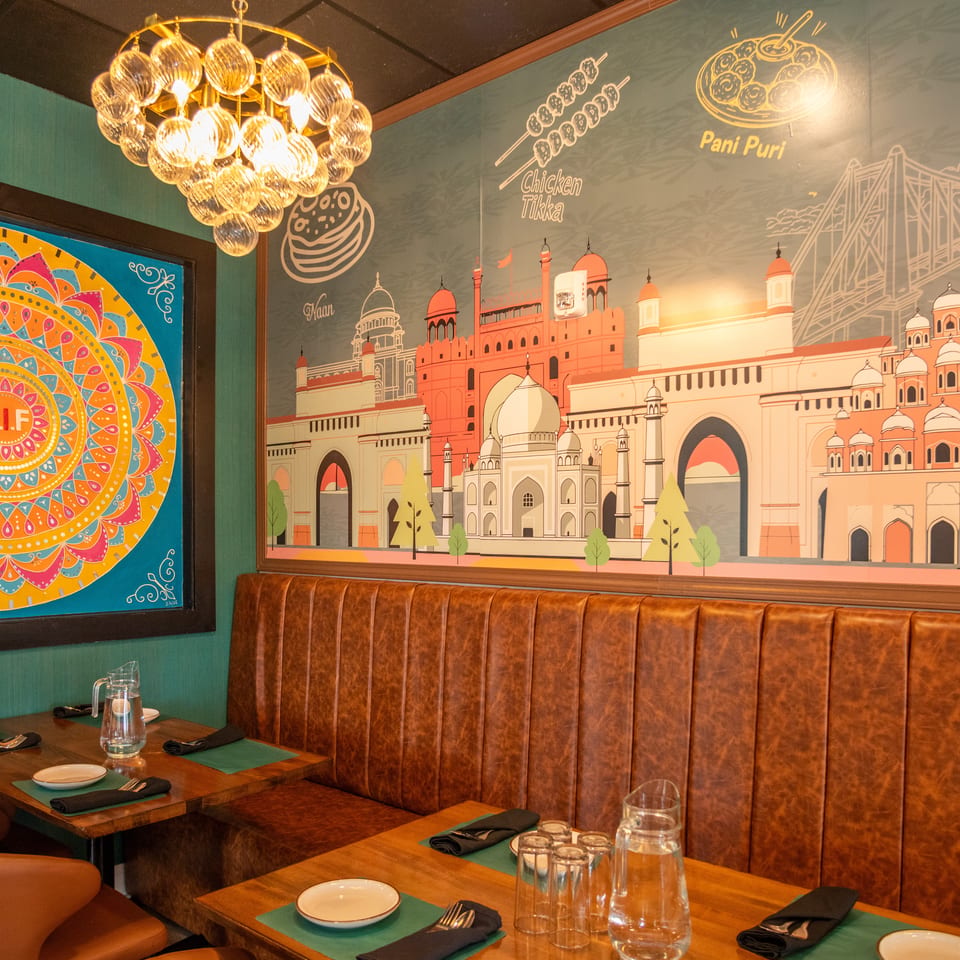 Restaurant interior with brown leather booths, wooden tables, and a colorful mural depicting Indian landmarks and food.