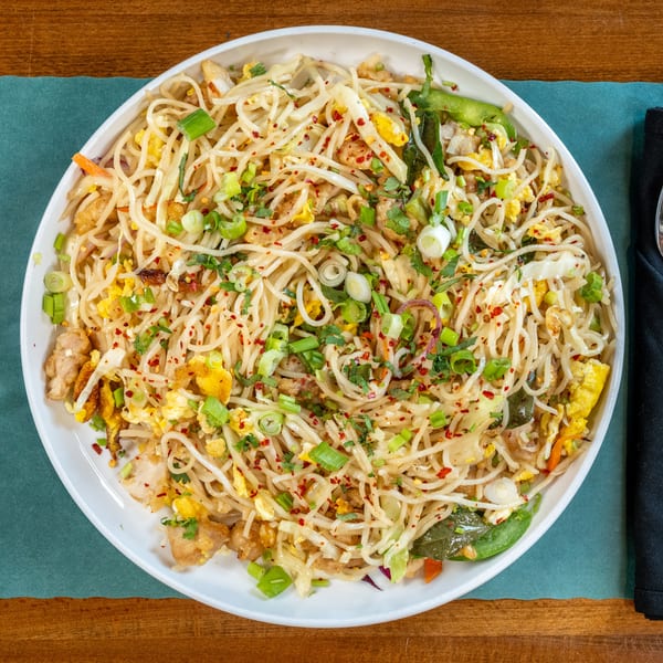 A white bowl of chicken chow mein with noodles, vegetables, and scrambled egg on a wooden table.
